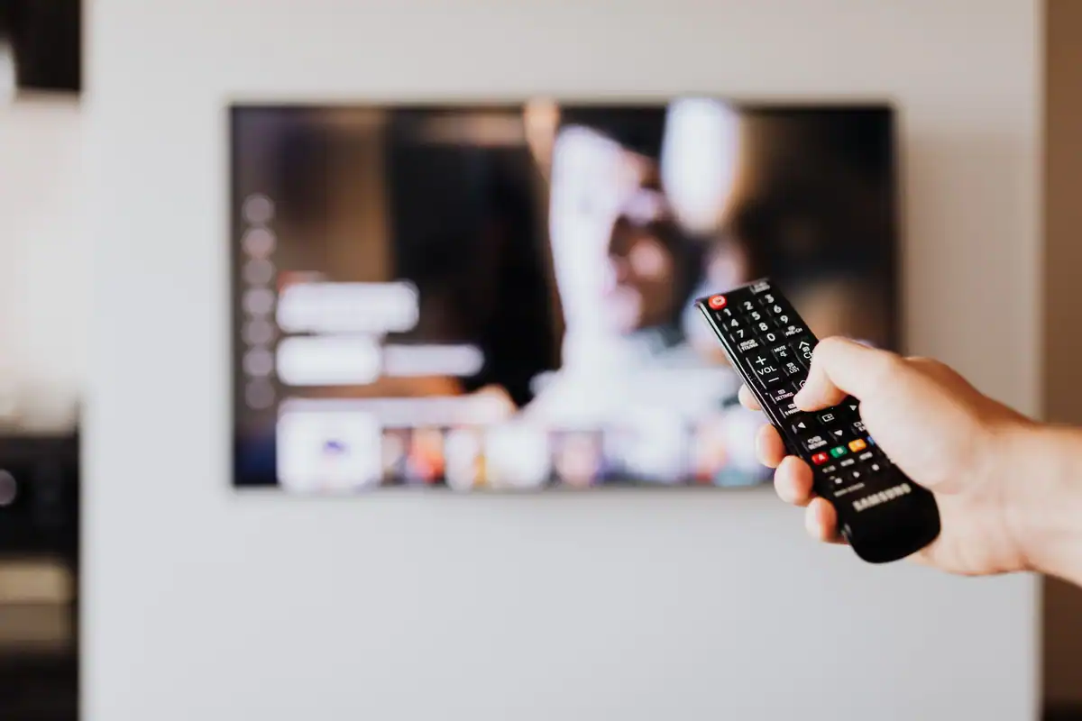 Persoană care folosește telecomanda, cu televizorul în fundal, aparat care consumă în standby